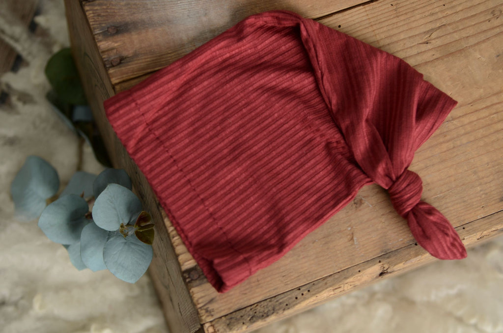 Flatlay image of Ribbed knot sleepy cap newborn photography prop in deep burgundy, styled on a rustic wooden bowl.
