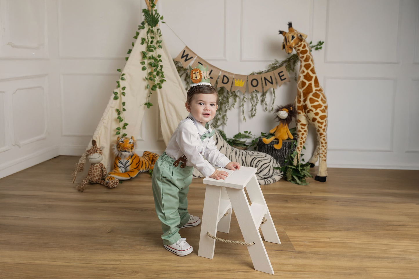 A jungle-themed first birthday setup featuring a toddler in suspenders standing beside a white wooden step ladder. Plush safari animals, a teepee with greenery, and a "Wild One" banner create a playful scene.