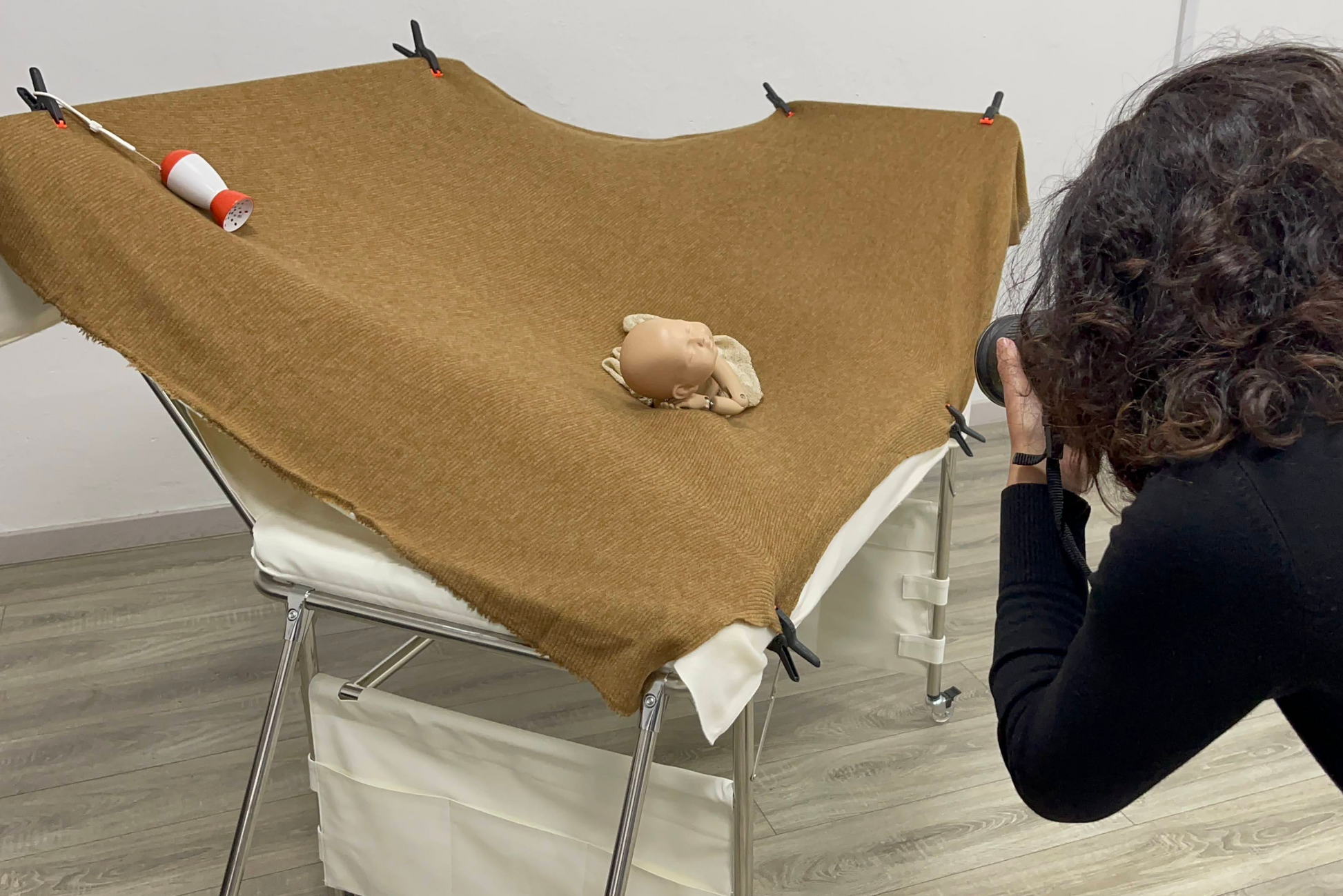 A baby photographer captures an image of a baby doll posed on an extended backdrop surface of a Newborn Photography Posing Table. The table is covered with a brown fabric that is secured with clips, showcasing the ample space for various posing options. The setup highlights the functionality and stability of the posing table for professional newborn photography.
