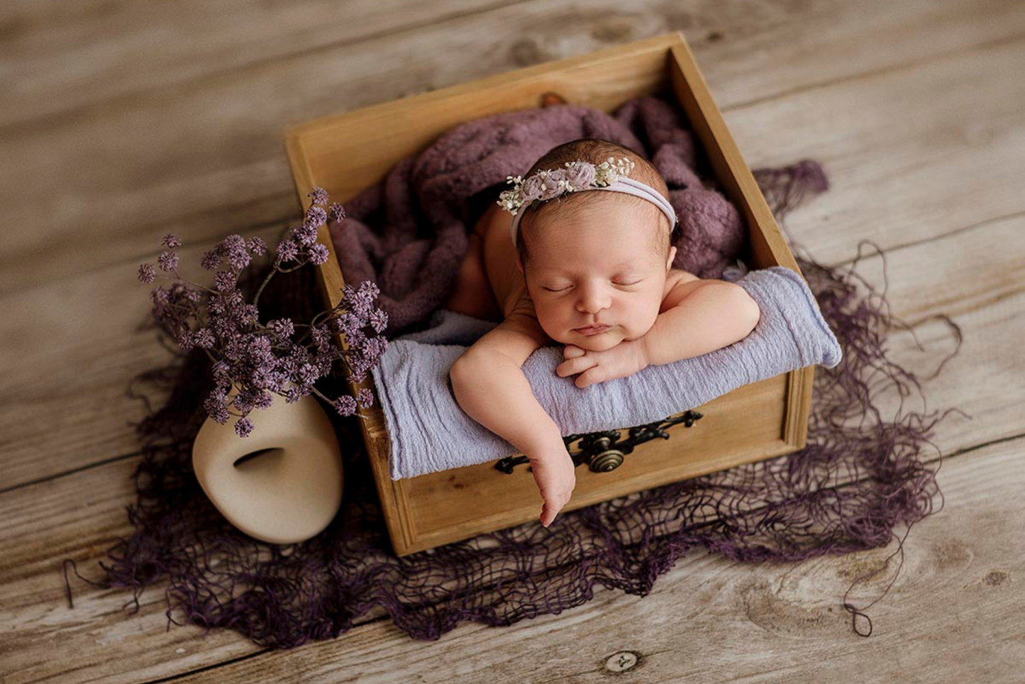 Rustic wooden box prop adorned with an ornate black latch, accompanied by delicate white fluff and a trailing green plant, set against a textured white backdrop. Perfect for newborn photography sessions. Available at Newborn Studio Props.