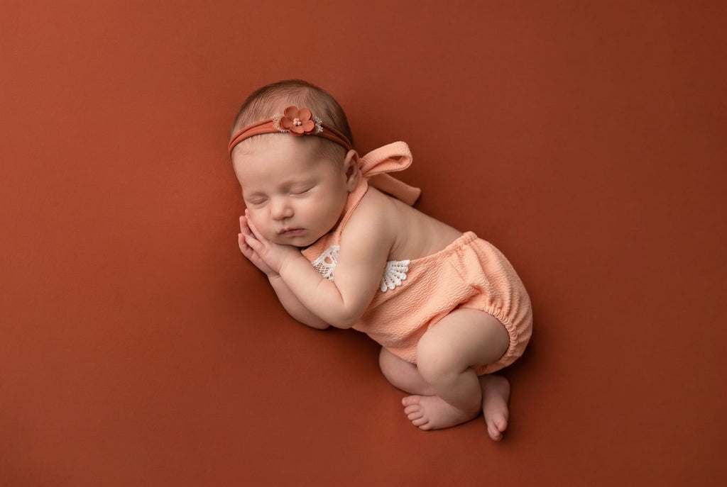 Newborn baby in peach outfit and headband on a brown background