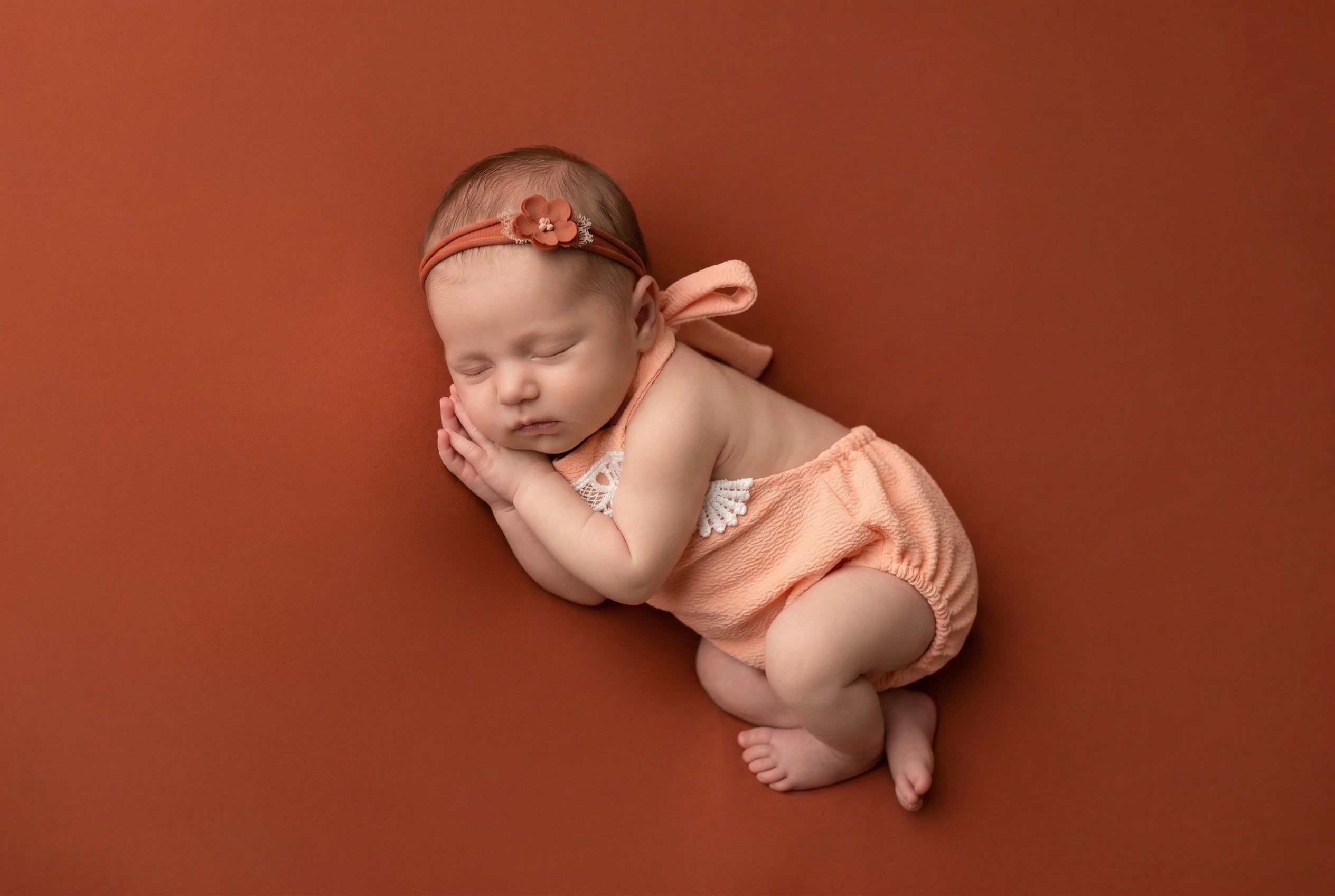 Newborn baby in peach outfit and headband on a brown background
