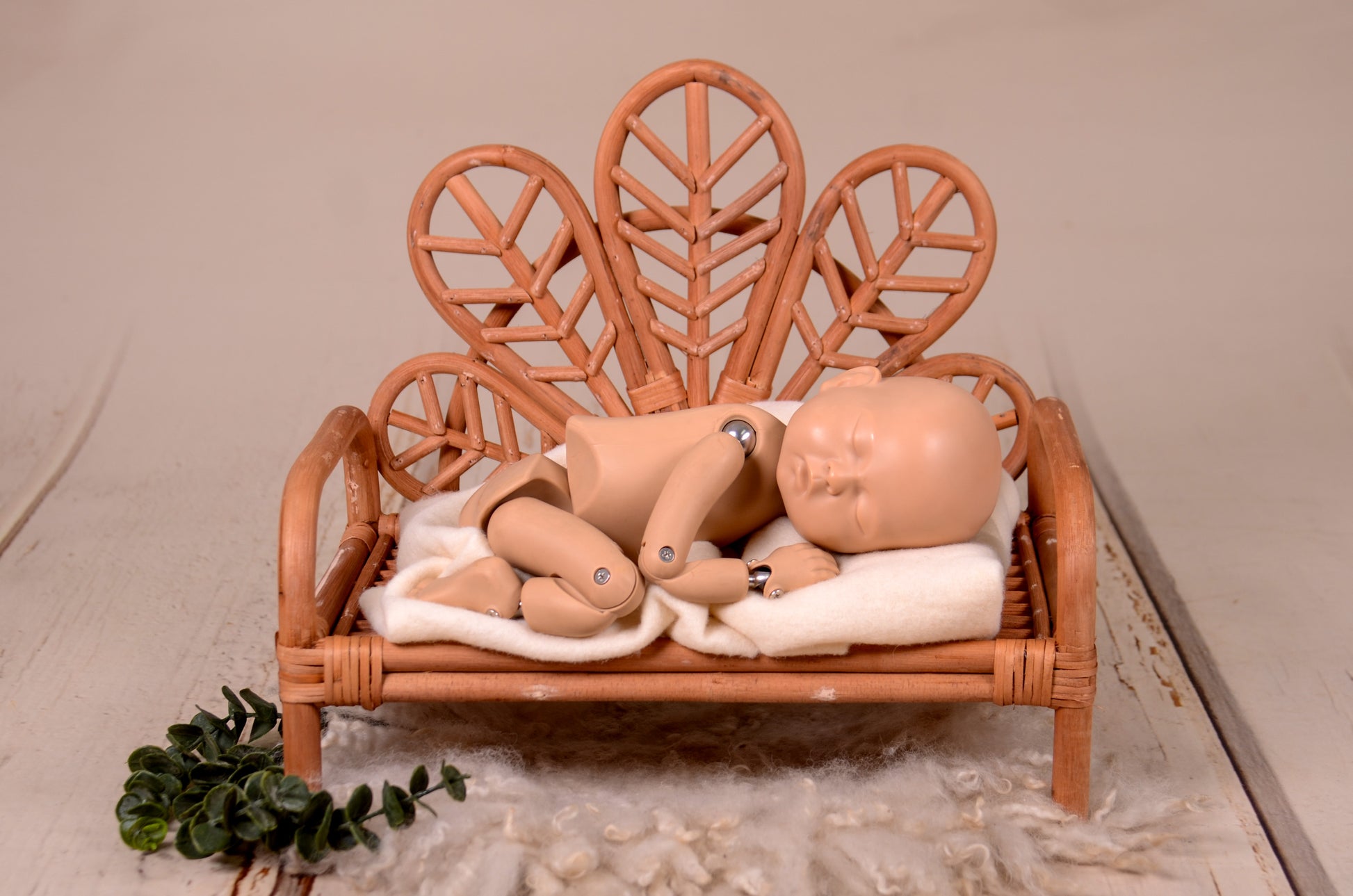 Handcrafted rattan bench with intricately woven leaf-shaped backrest, showcased on a distressed white wooden backdrop with soft faux fur and decorative greenery at the base. Ideal prop for newborn photography sessions, available exclusively at https://newbornstudioprops.com/