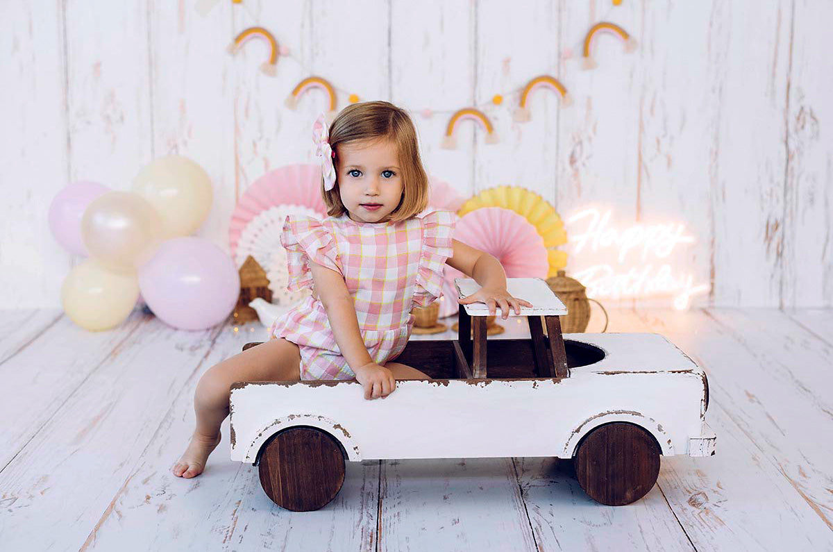 Toddler in plaid dress on a white-brown truck prop with a pastel birthday backdrop.