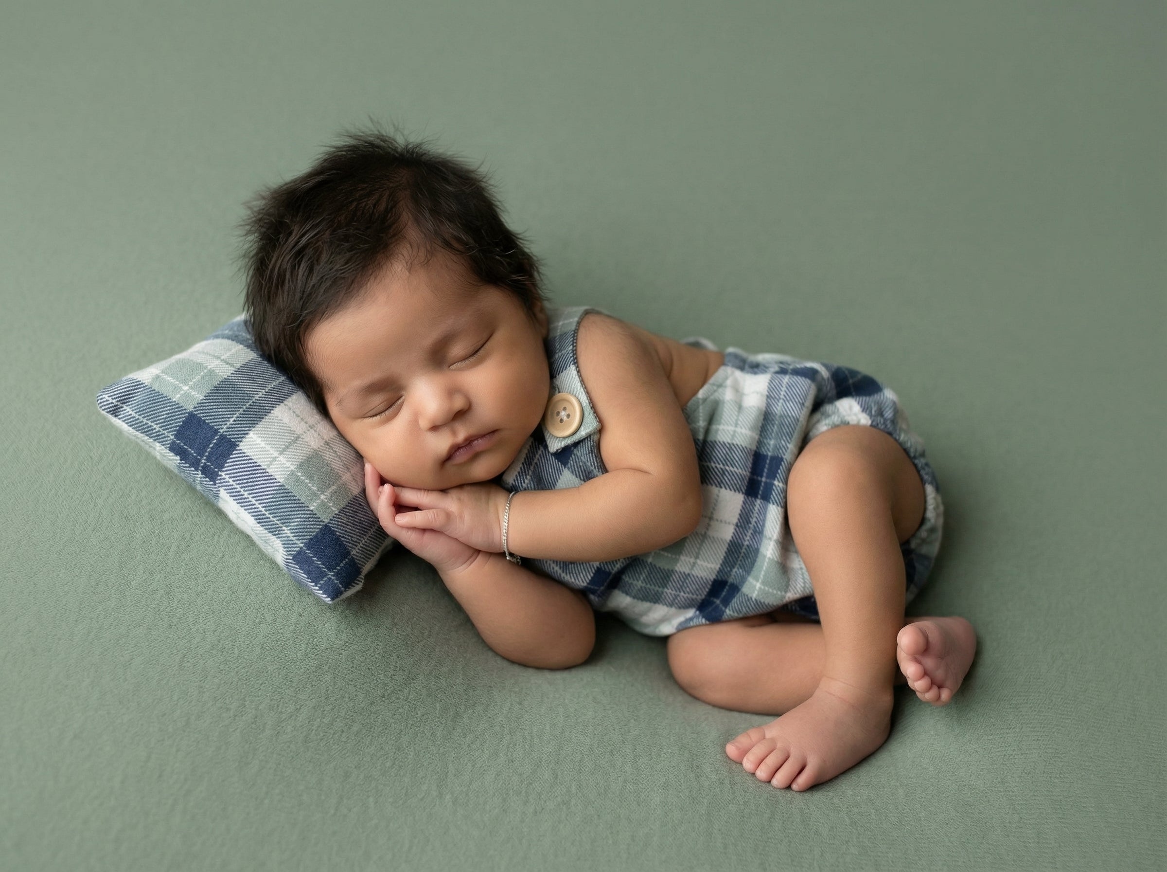 Sleeping baby in blue plaid romper on matching pillow, newborn photography prop set on soft sage backdrop.