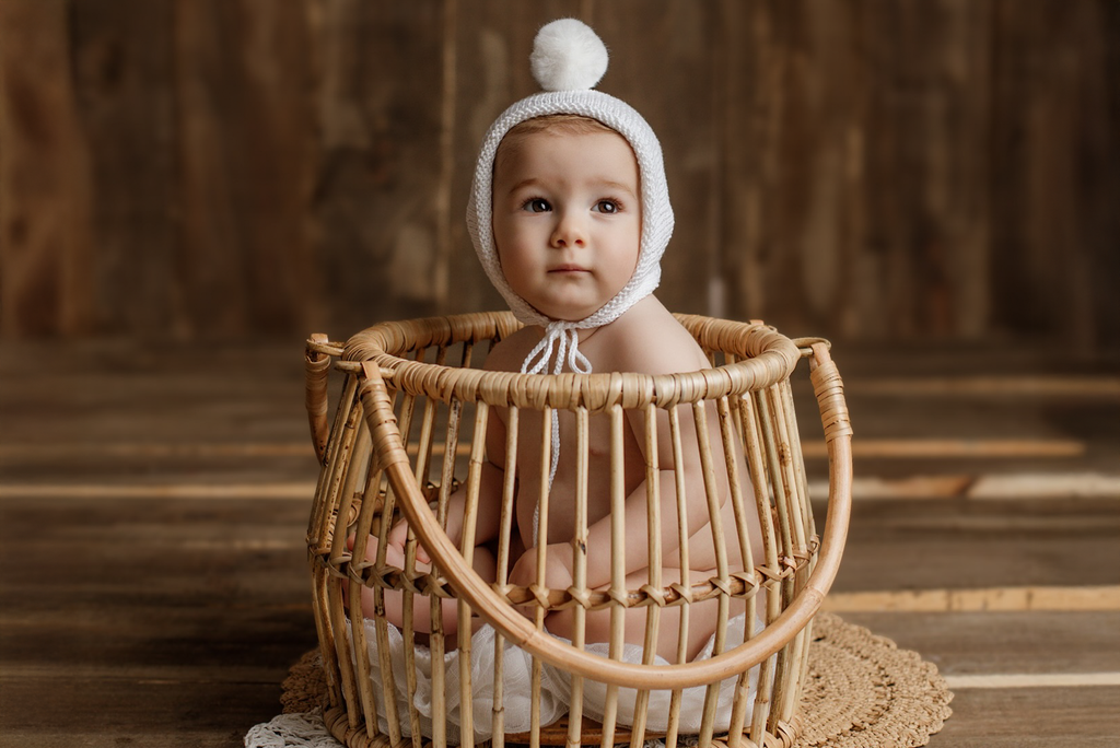 Adorable baby sits inside a "Woven Wonders" rattan basket, wearing a white knit bonnet with a pompom. The rustic wooden backdrop and cozy textures create a warm, vintage-inspired newborn photography prop setup.