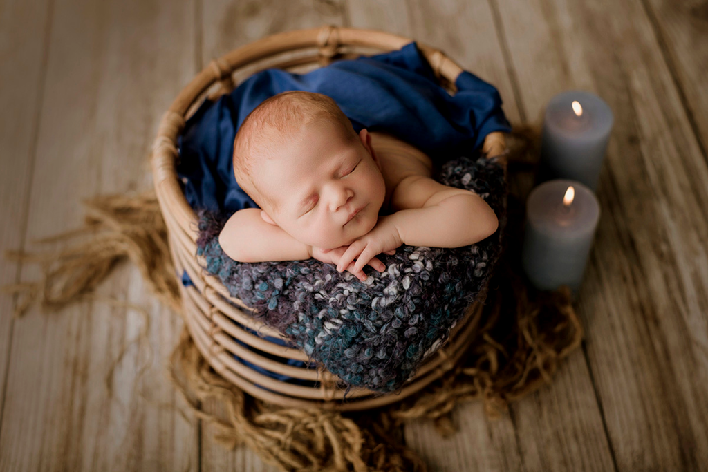 A beautifully handcrafted rustic rattan bowl serves as the perfect newborn photography prop, cradling a peacefully sleeping baby. The soft blue wrap adds warmth and texture, complementing the earthy tones of the wooden floor. The baby rests with tiny hands tucked under its chin, creating a serene and timeless portrait.