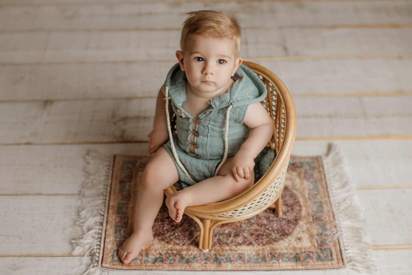 Adorable baby sits in a rattan nest cocoon, a charming baby and toddler photography prop, on a vintage rug.