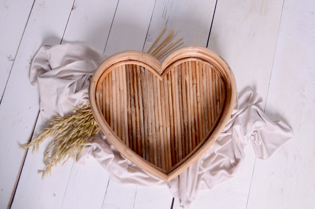 Overhead shot of the Rattan Heart Bowl on a white wooden floor, styled with soft fabric and dried wheat, presenting a timeless and elegant newborn photography setup.
