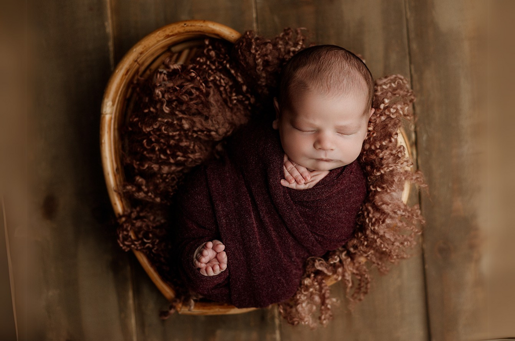 Newborn baby peacefully sleeping in the Rattan Heart Bowl, nestled in a cozy brown knit wrap with a matching fur layer, creating a warm and intimate newborn photography setup.