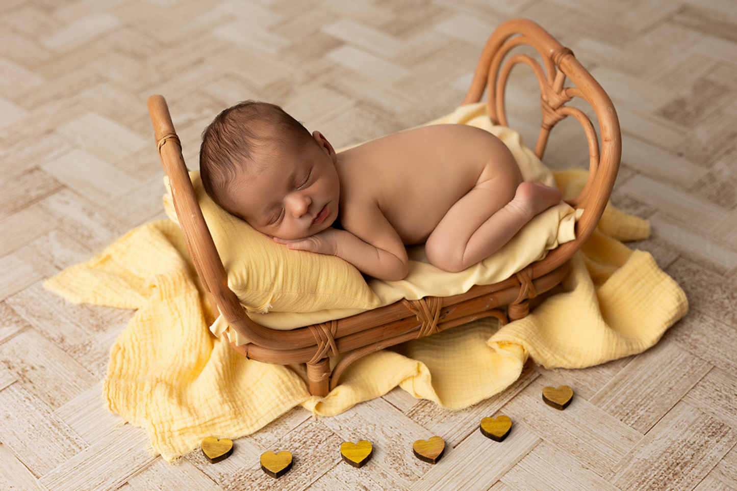 Sleeping baby cradled in rattan prop on yellow blanket, wooden floor setting.