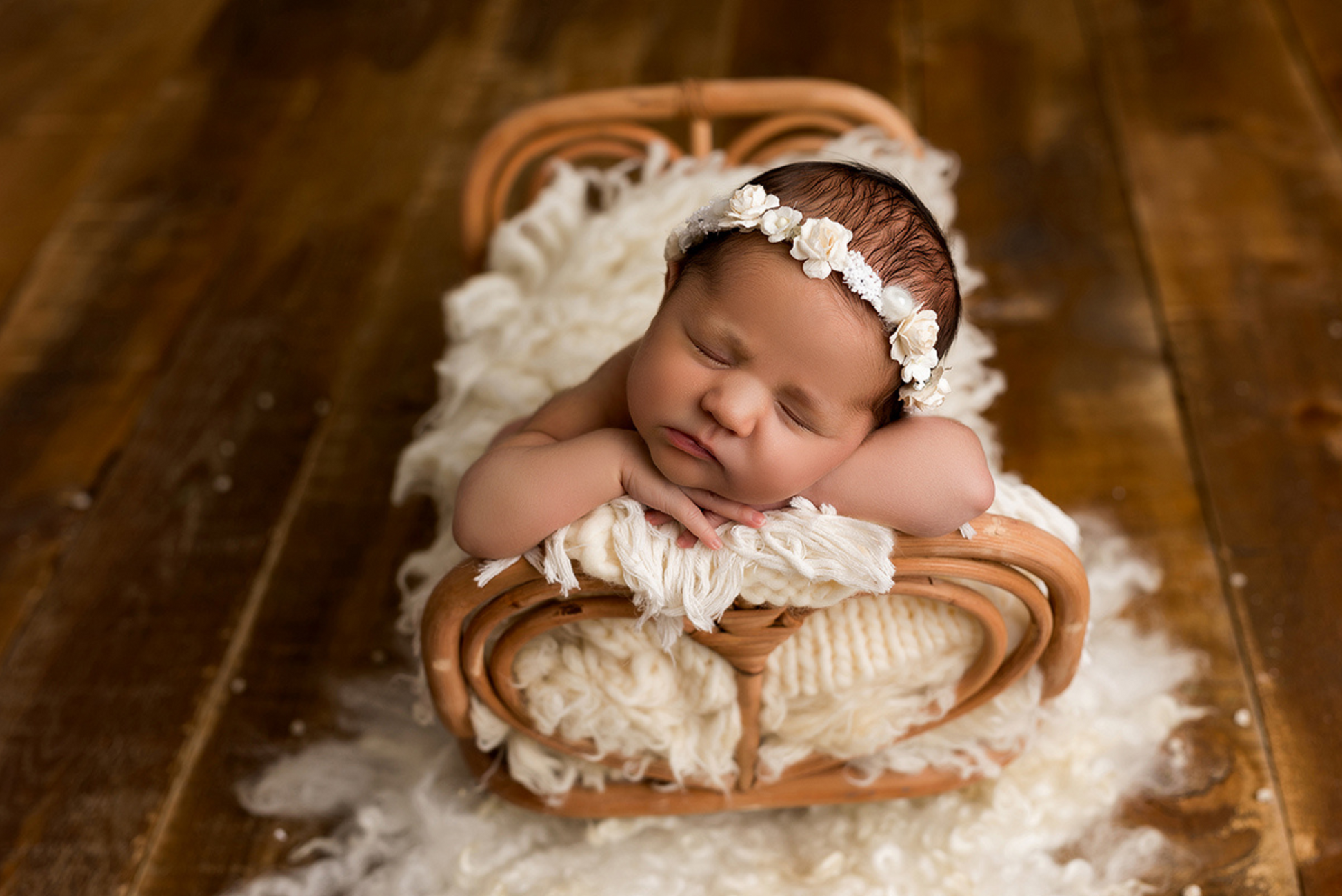 Newborn in rattan love nest bed, adorned with a delicate white headband, on rustic wood.