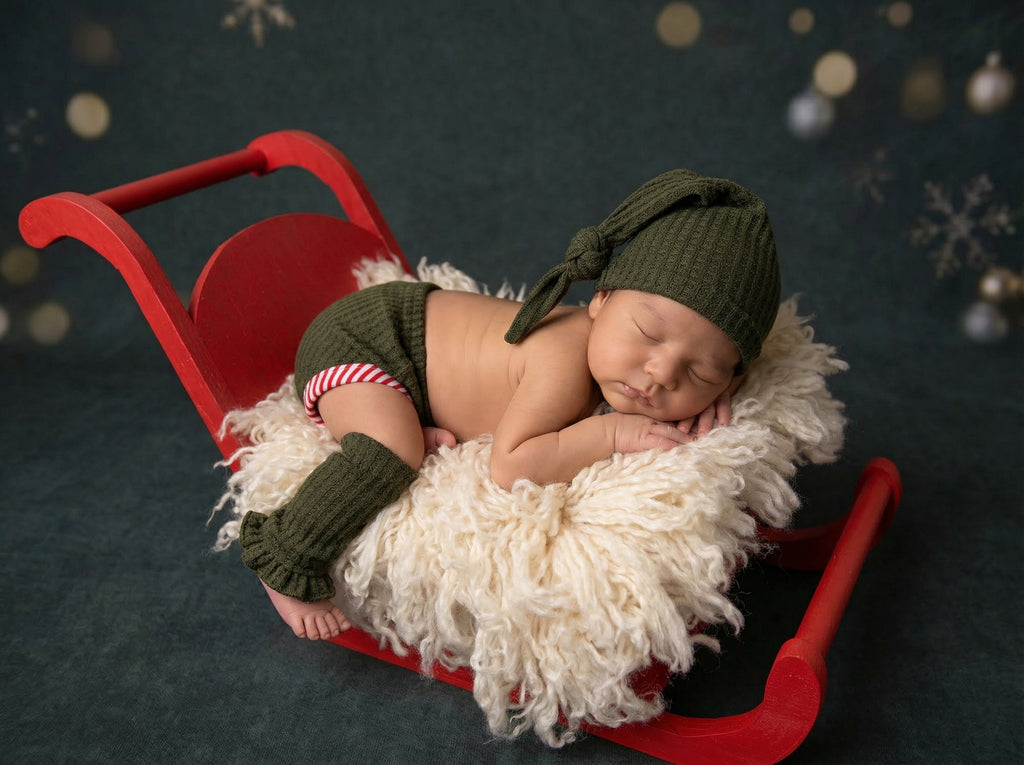 Newborn baby in green outfit, leg warmers, and hat lying on a red sleigh. Newborn photography Christmas clothing set