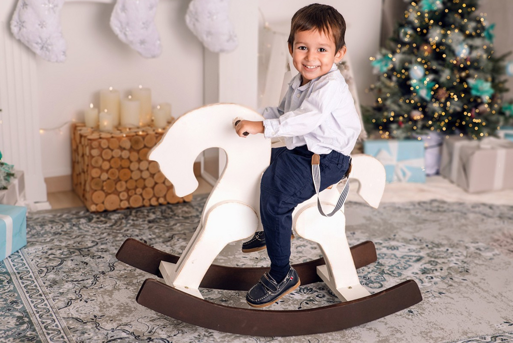 A smiling child seated on a white wooden rocking horse in a cozy Christmas setting with a tree, stockings, and candles. Perfect for holiday-themed newborn photography props.