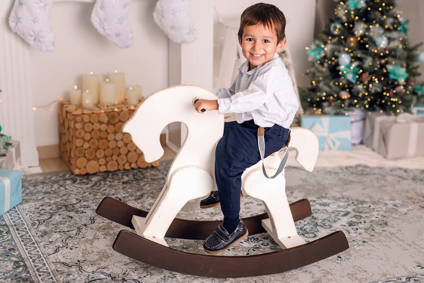 A smiling child seated on a white wooden rocking horse in a cozy Christmas setting with a tree, stockings, and candles. Perfect for holiday-themed newborn photography props.