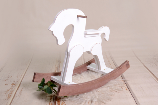 A white wooden rocking horse with natural wood rockers, styled on a cream backdrop with a fluffy rug and greenery. Ideal as a newborn photography prop.