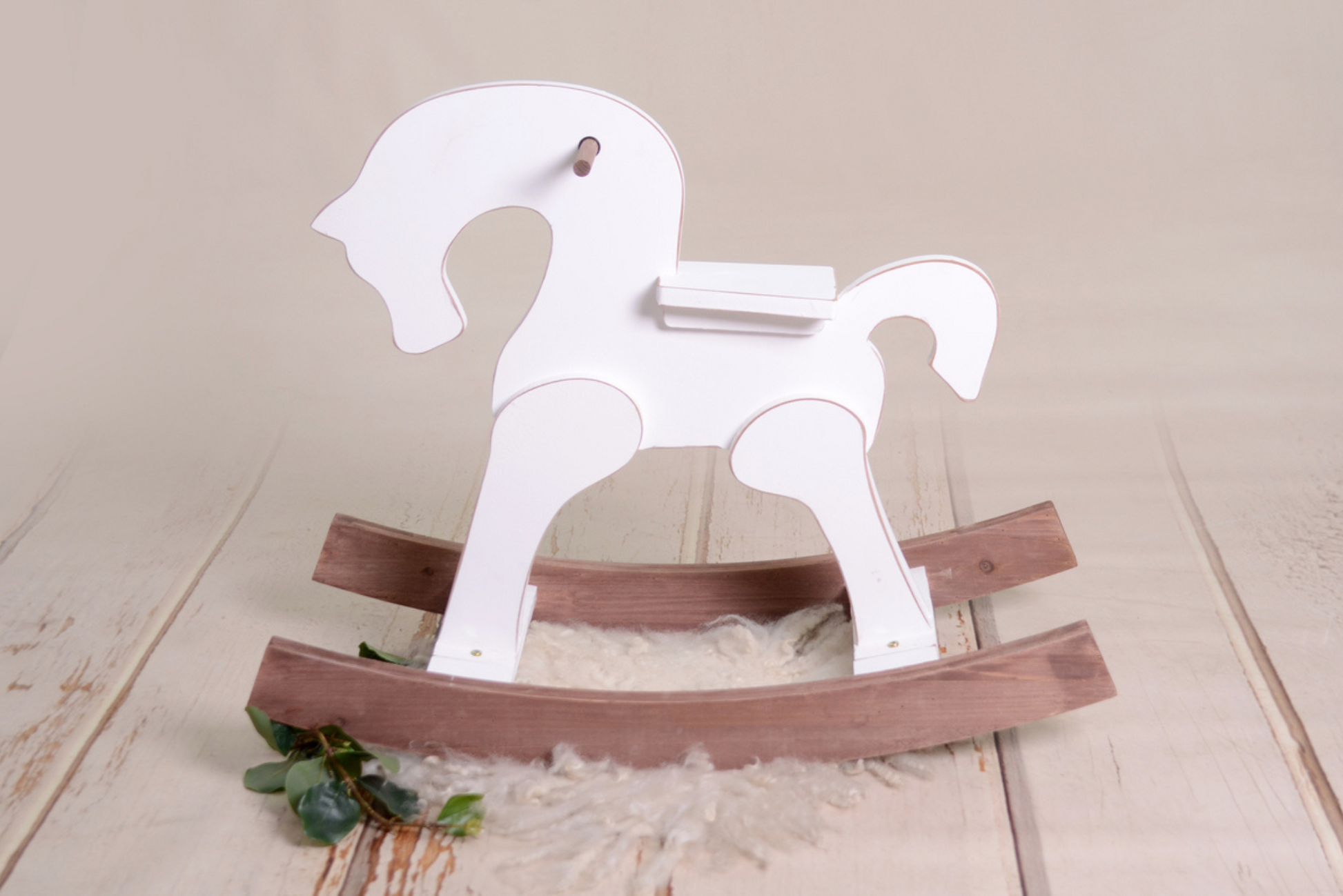 
A charming white wooden rocking horse with natural-toned rockers, set against a soft cream backdrop adorned with a cozy rug and a touch of greenery. Perfectly suited as a timeless prop for newborn photography sessions.