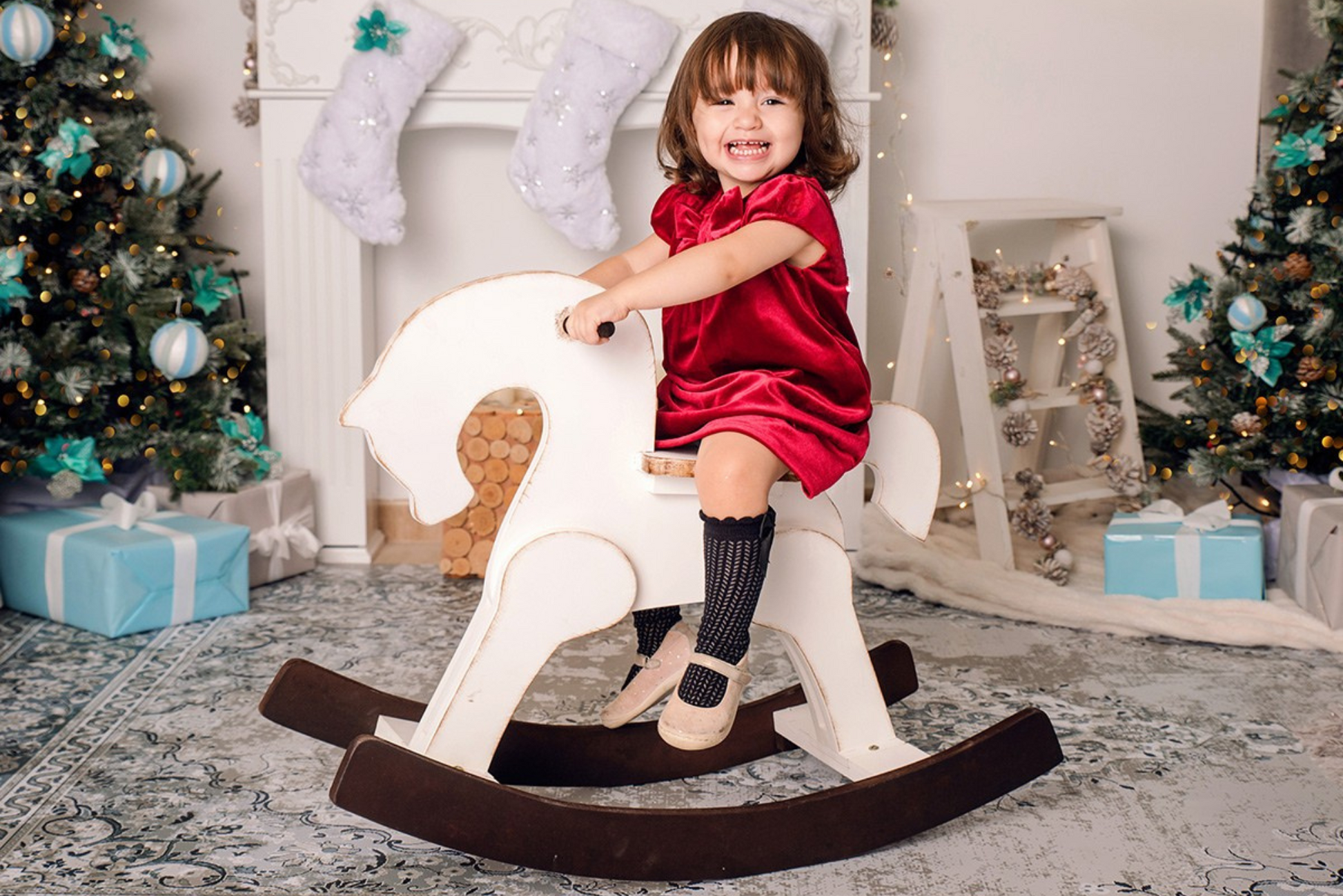 A cheerful child in a festive red velvet dress is enjoying a ride on a white wooden rocking horse with dark brown rockers. The scene is set in a beautifully decorated holiday backdrop featuring Christmas trees, stockings, and wrapped presents, creating a warm and magical atmosphere for photography.