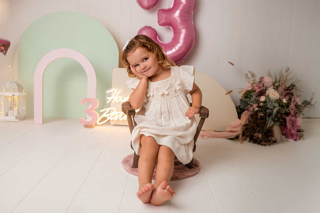Toddler in cream dress poses on brown Wooden Orbit Chair, a charming newborn photography prop.