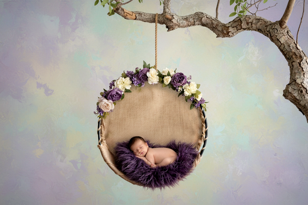 Newborn photography prop featuring a round burlap swing with purple florals and fluffy violet bedding.