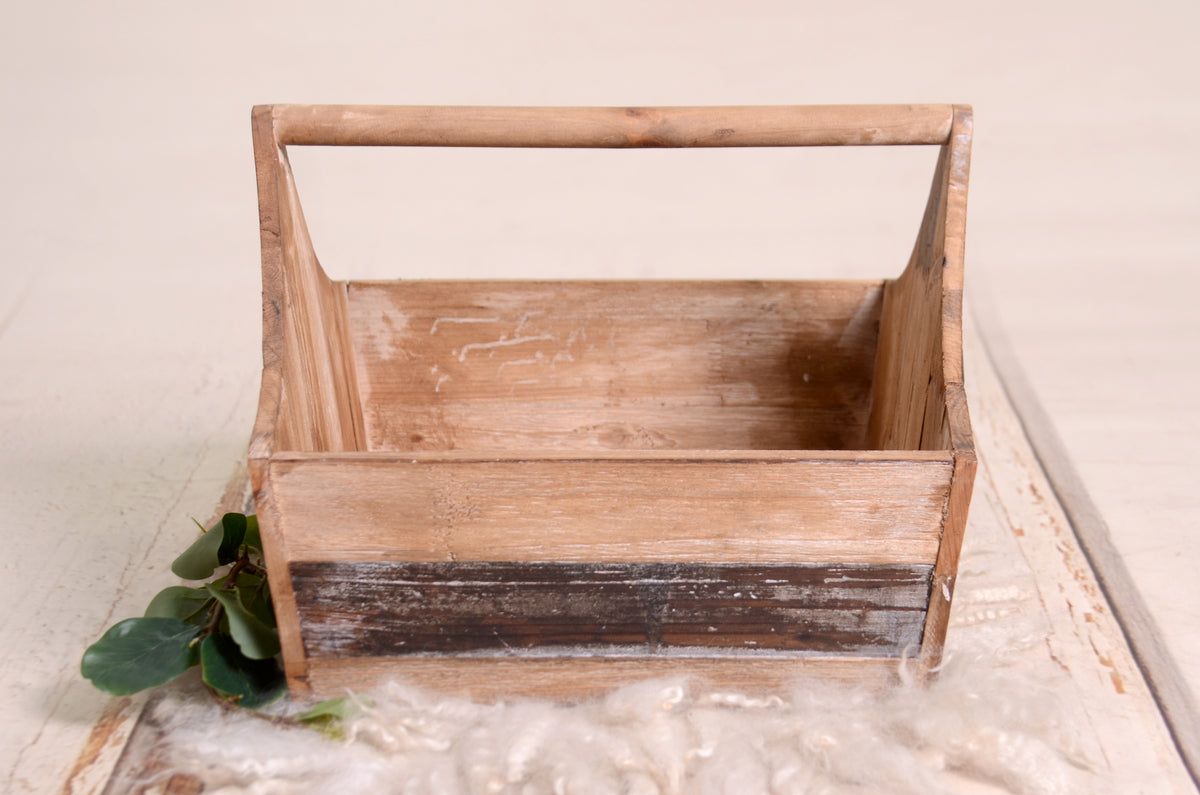 Handcrafted wooden basket with a curved handle, styled on a textured wool layer and greenery for newborn photography.