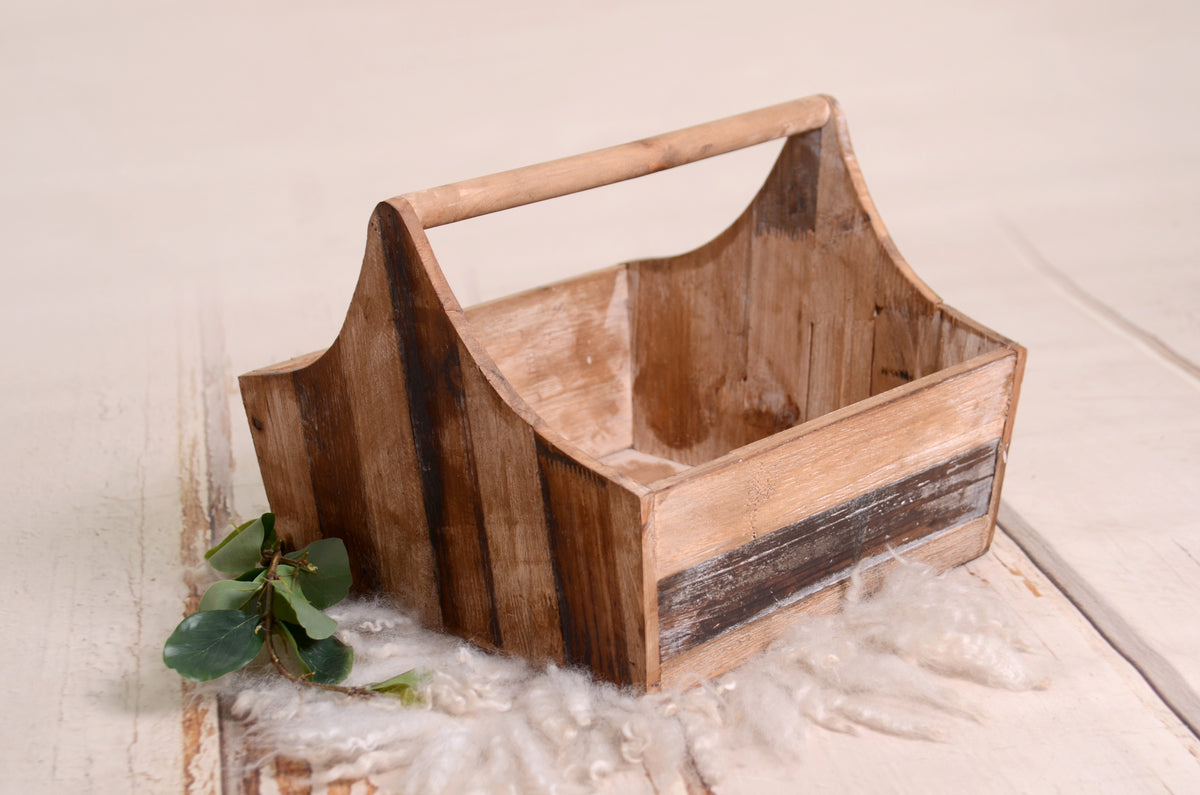 Rustic wooden basket with arched handle, natural finish, accented by greenery and soft wool; ideal for newborn photography props.