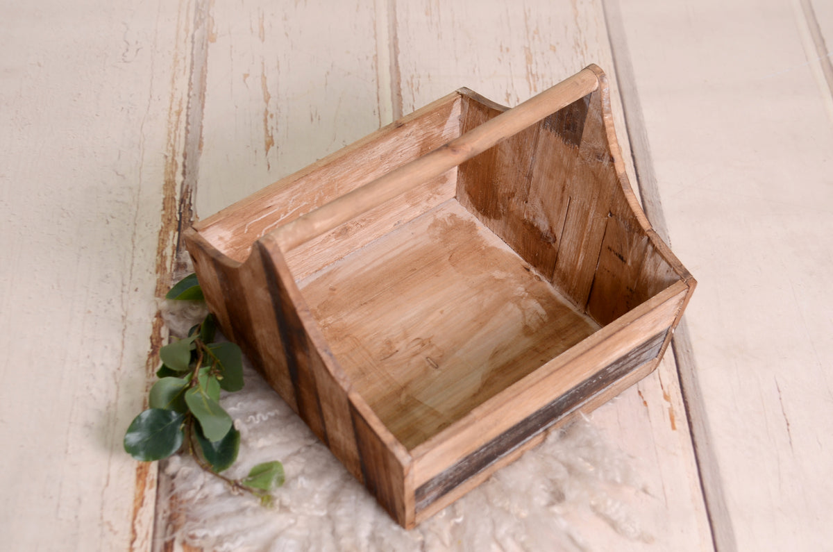 Vintage-inspired wooden basket with arched handle, placed on soft wool and accented with greenery; a charming newborn photography prop.