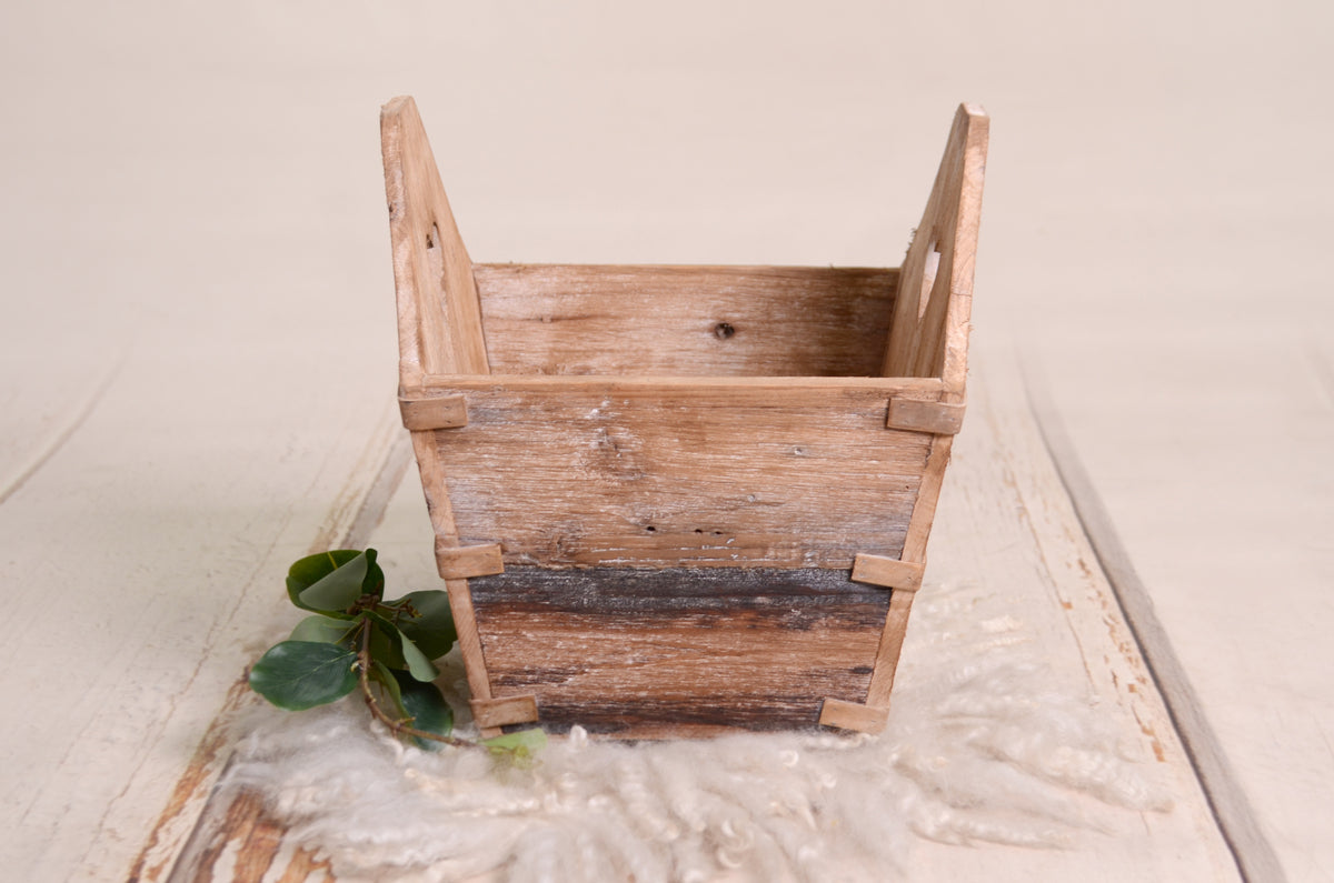 A weathered wooden crate with heart-shaped handles, perfect as a newborn photography prop. Styled with wool fabric and greenery for a cozy, rustic touch.