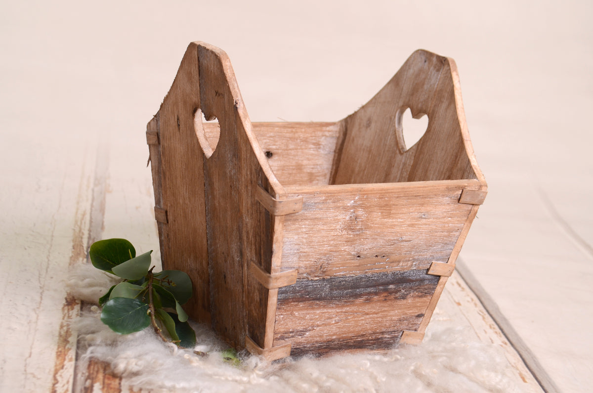 A rustic wooden crate with heart-shaped cutouts, ideal as a newborn photography prop. Set on a soft wool layer with greenery accents, the crate adds a vintage charm to photography setups.