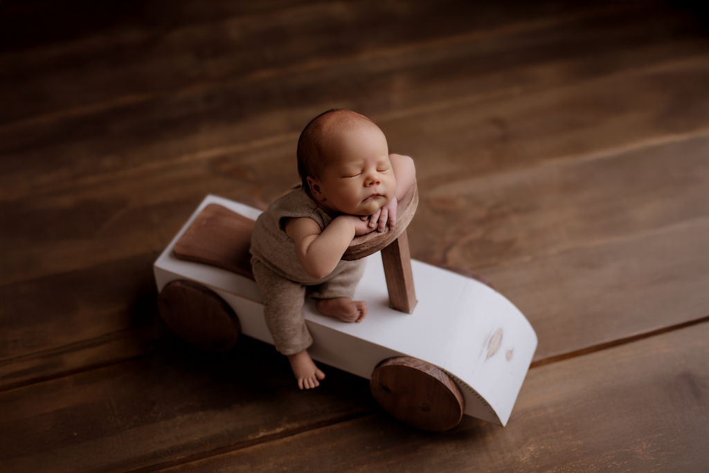 Newborn posed on Rustic Ride-On, a wooden car-shaped newborn photography prop, with warm wood floor.
