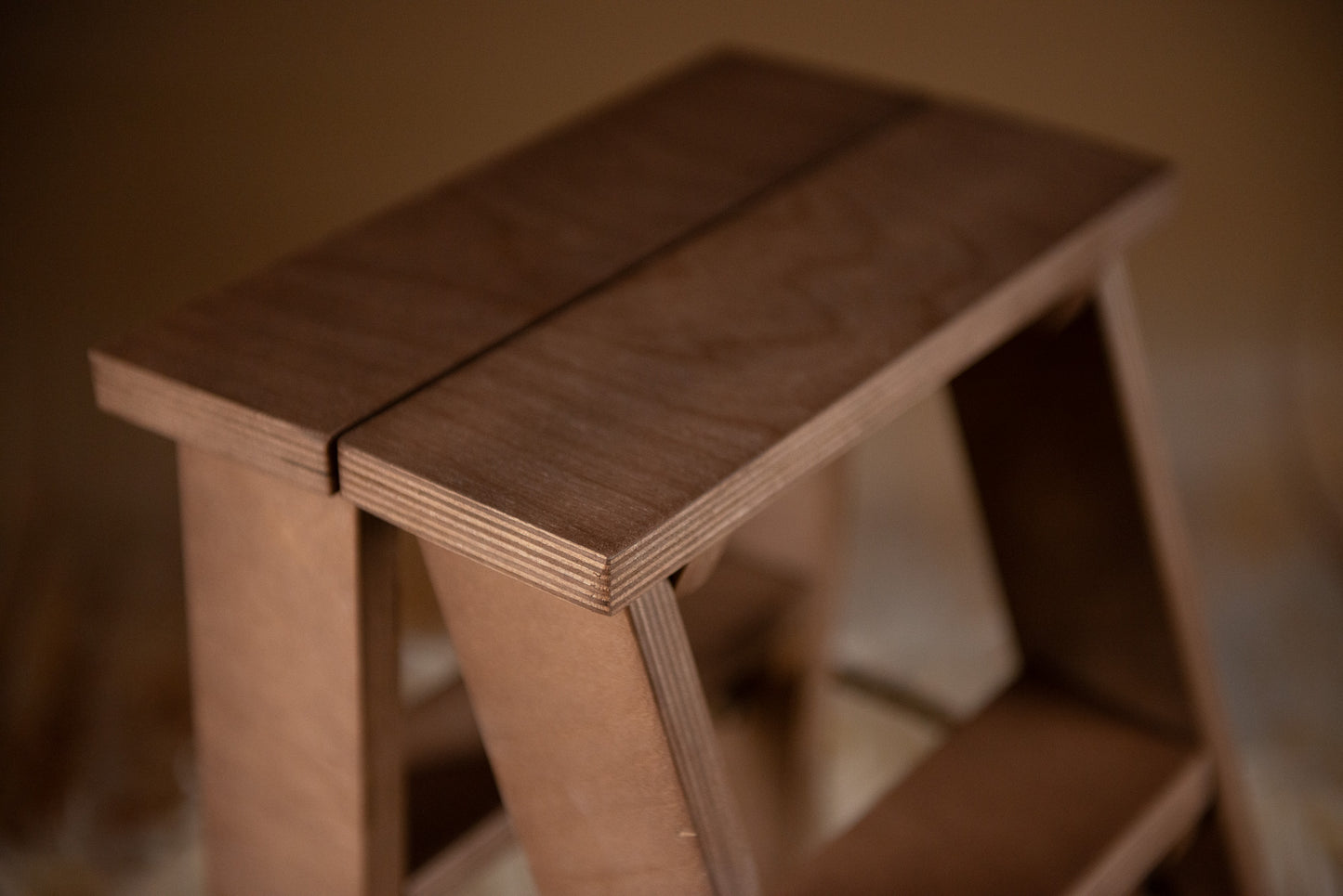 Kid Photography Prop. A small, wooden step ladder with a natural finish is placed on a fluffy, beige faux fur rug. The step ladder has two steps and a rope handle on the side for easy carrying. The background features a warm, brown gradient, creating a cozy and rustic ambiance.