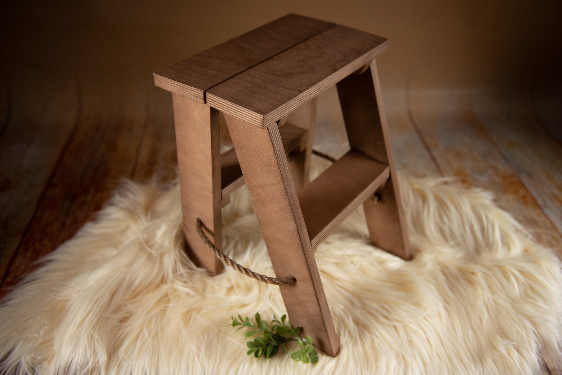 Toddler Photography Prop. A small, wooden step ladder with a natural finish is placed on a fluffy, beige faux fur rug. The step ladder has two steps and a rope handle on the side for easy carrying. The background features a warm, brown gradient, creating a cozy and rustic ambiance.