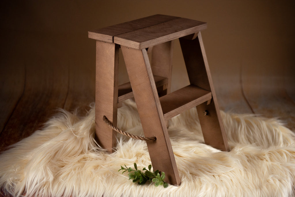 Kid Photography Prop. A small, wooden step ladder with a natural finish is placed on a fluffy, beige faux fur rug. The step ladder has two steps and a rope handle on the side for easy carrying. The background features a warm, brown gradient, creating a cozy and rustic ambiance.