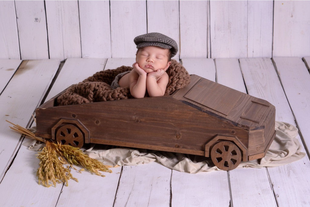 Newborn photography prop styled as a wooden Tesla Cybertruck with baby in cap and cozy brown blanket.