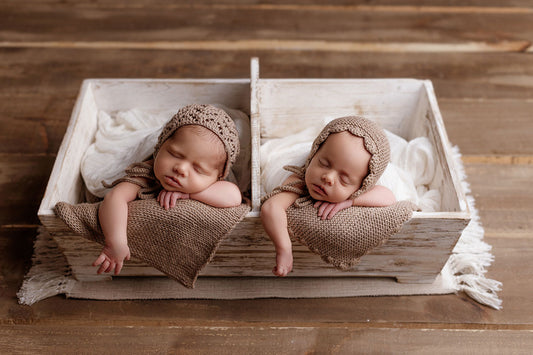 wo newborn babies peacefully sleeping in a rustic white wooden twin crate, their tiny arms resting over the edge. The crate is lined with soft fabric, creating a cozy setting on a wooden floor.