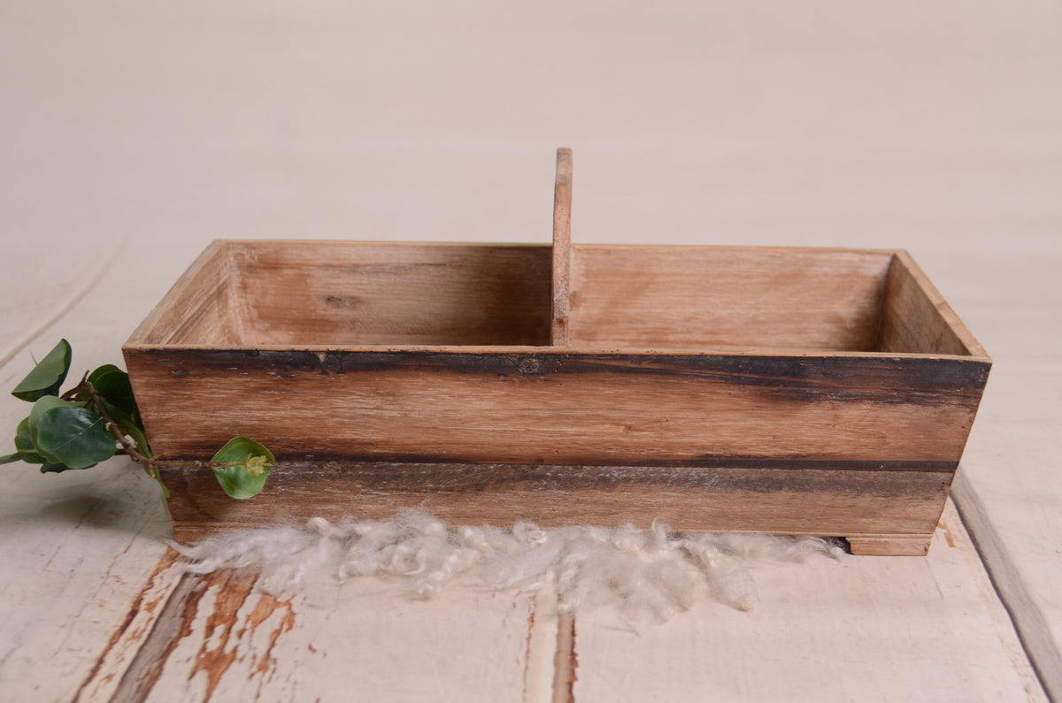 Rustic twin snuggle crate in distressed brown wood, designed as a newborn photography prop. Features a central divider for twin posing, with a vintage charm. Set on a white wooden floor with soft wool and greenery for a warm, organic feel.