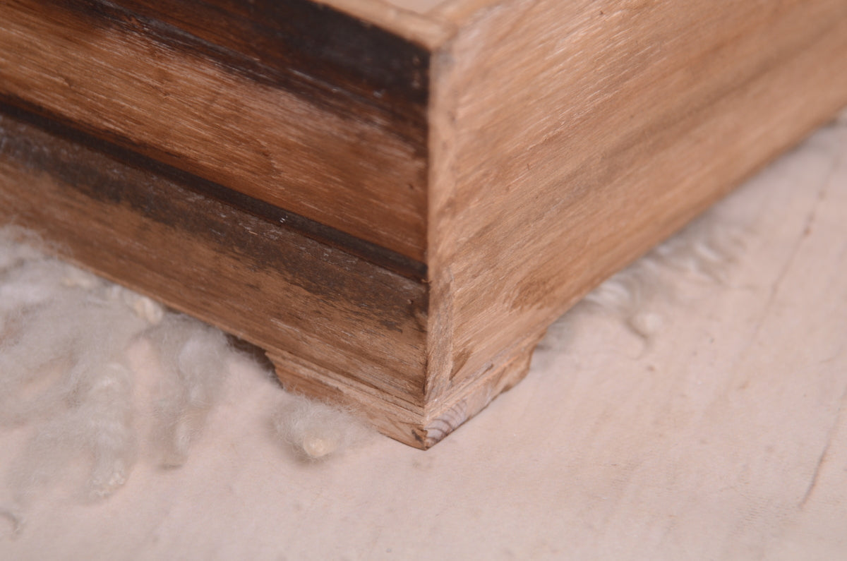 Close-up of a rustic wooden twin snuggle crate, a perfect newborn photography prop. The distressed brown finish and sturdy frame add vintage charm. Captured from a corner angle, highlighting the texture and craftsmanship, set against a white wooden floor with soft wool and greenery for a natural touch.