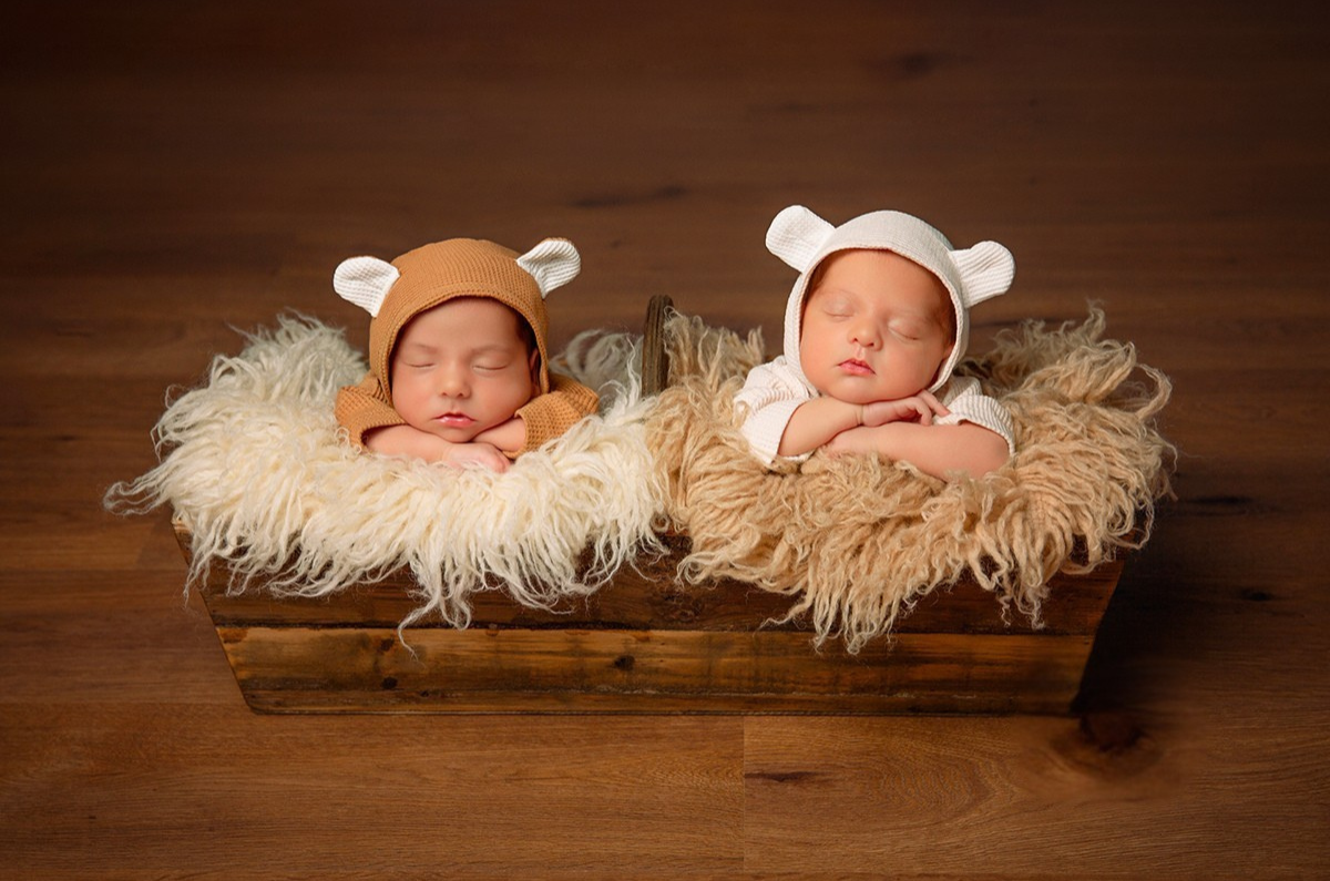 Twin newborns sleep peacefully in a rustic wooden crate, nestled in fluffy faux fur. They wear matching bear-ear bonnets, one in warm brown and the other in soft cream. This cozy **newborn photography prop** setup creates a heartwarming and serene scene.