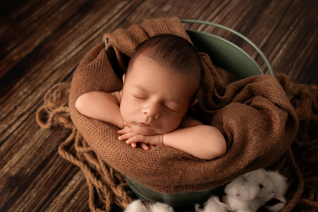 Newborn photography prop setup with baby in green vintage metal bucket, wrapped in soft brown knit.