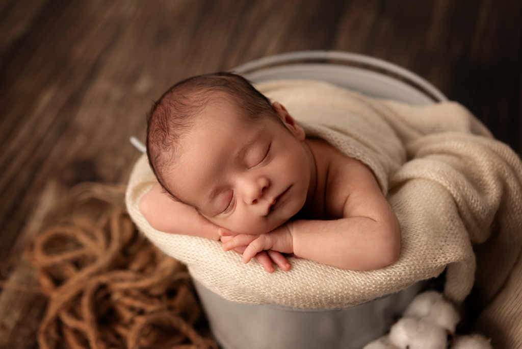 Newborn photography prop of baby sleeping in vintage metallic bucket with cream knit blanket wrap.