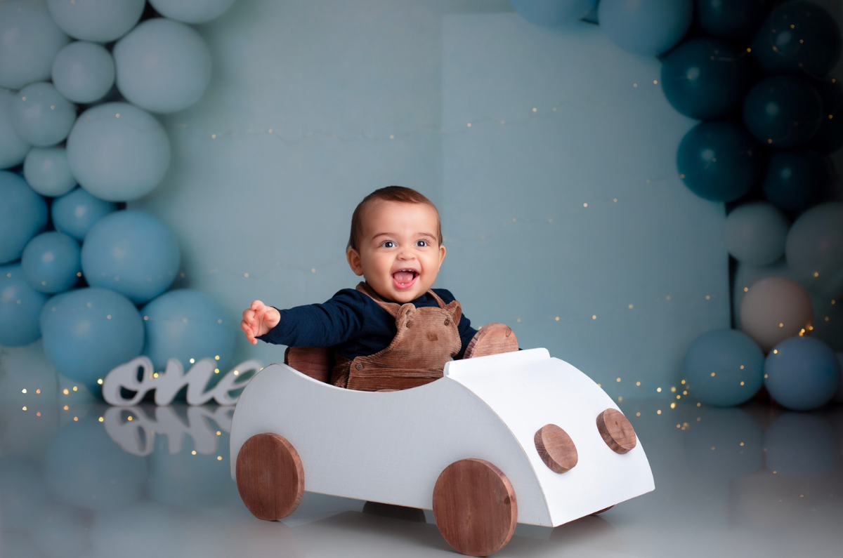 Smiling baby boy sits in a white wooden car, a stylish newborn photography prop. The setup includes a dreamy blue balloon backdrop with soft fairy lights, creating a joyful and celebratory atmosphere.