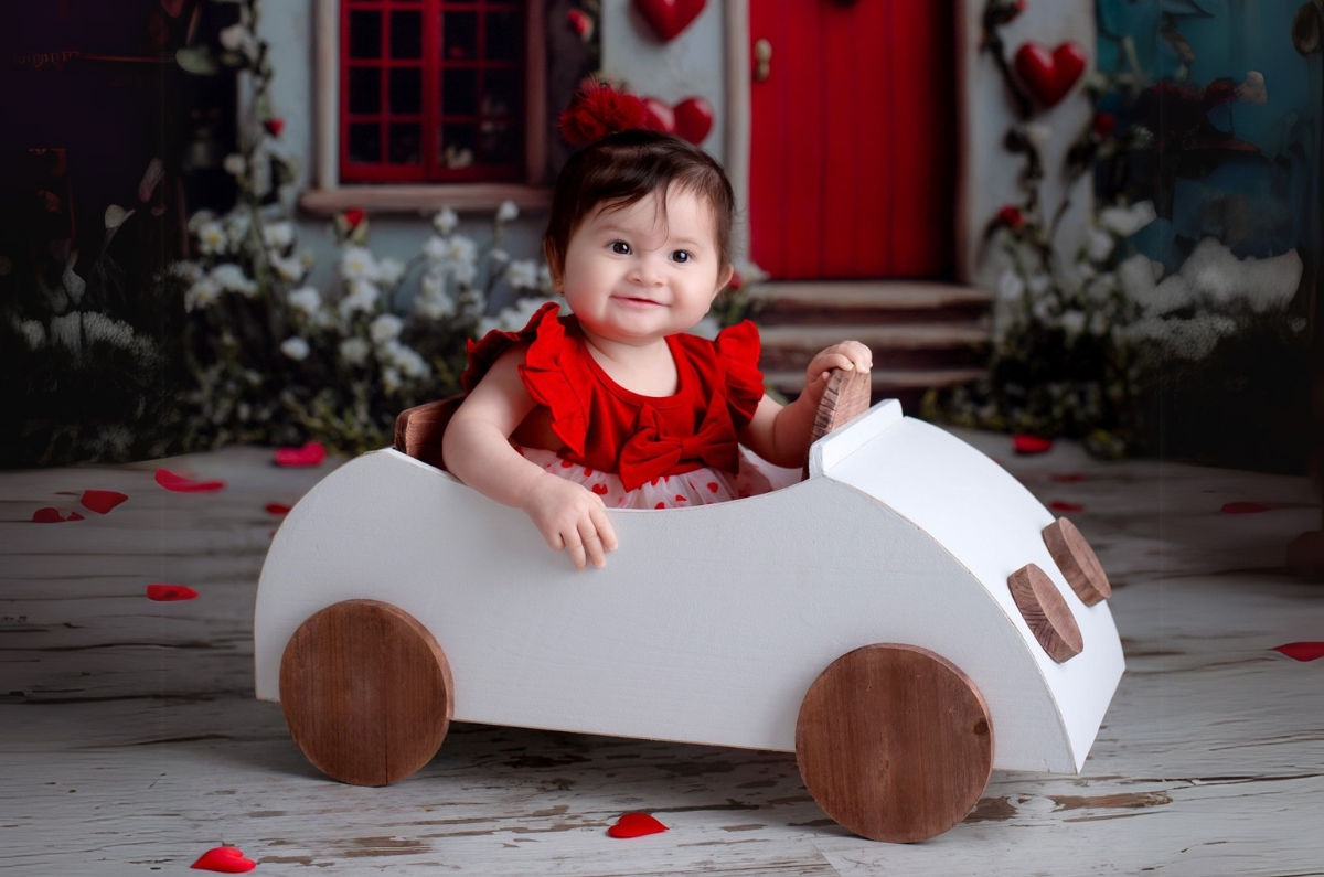 Adorable baby girl in a red dress sits in a white wooden car, a charming newborn photography prop. The setup features a romantic backdrop with red hearts, flowers, and a cozy cottage door, creating a warm and festive scene.