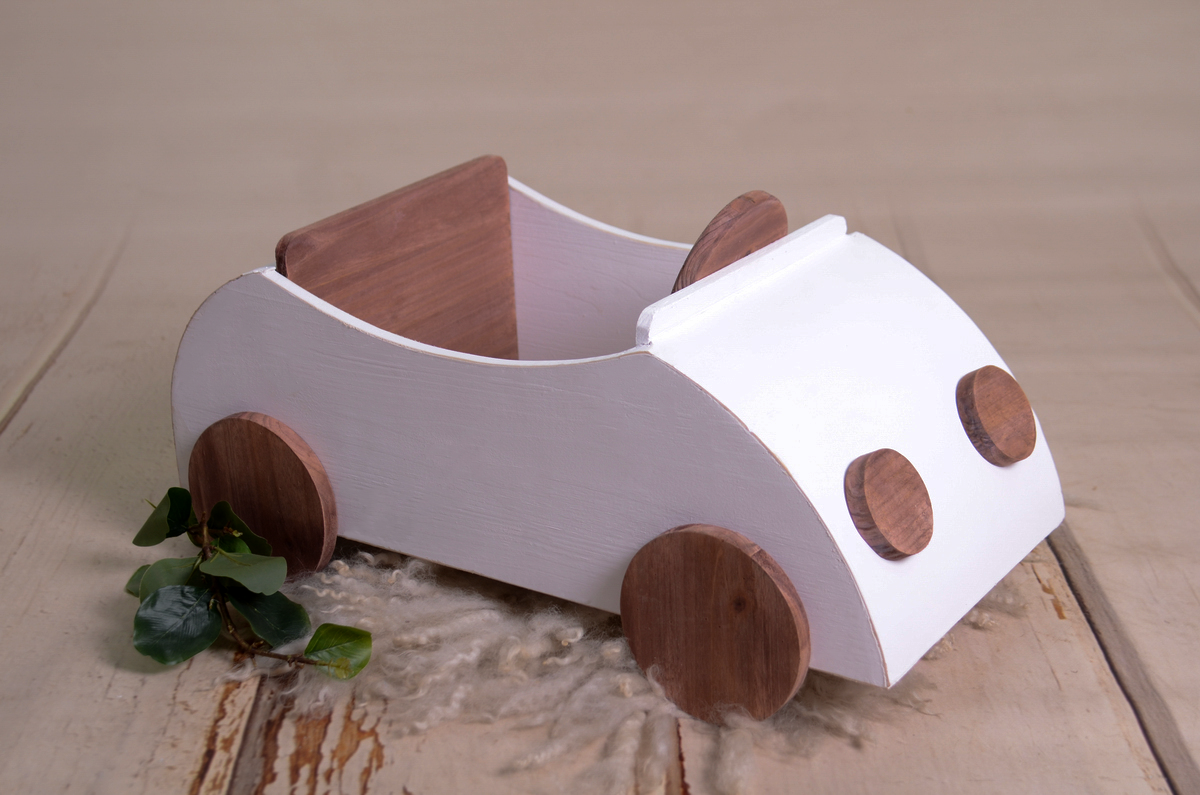 Full view of a handcrafted wooden VW Beetle cabriolet newborn photography prop. The car-shaped prop has a white-painted body with natural wood wheels, headlights, and a tiny steering wheel, placed on a rustic wooden floor with soft fabric underneath.