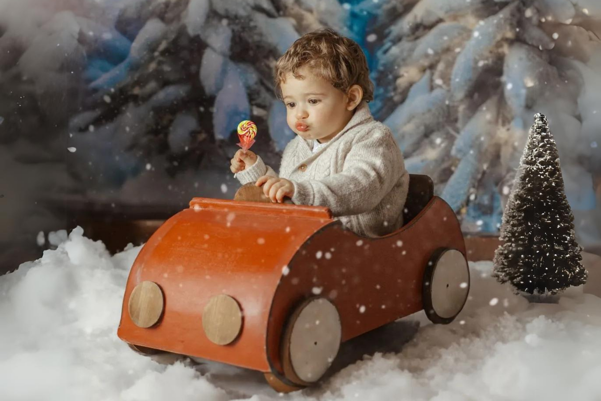 A toddler sits in a red wooden toy car, surrounded by snowy scenery, holding a colorful lollipop. A small, frosted tree adds to the wintery theme. Newborn photography prop.