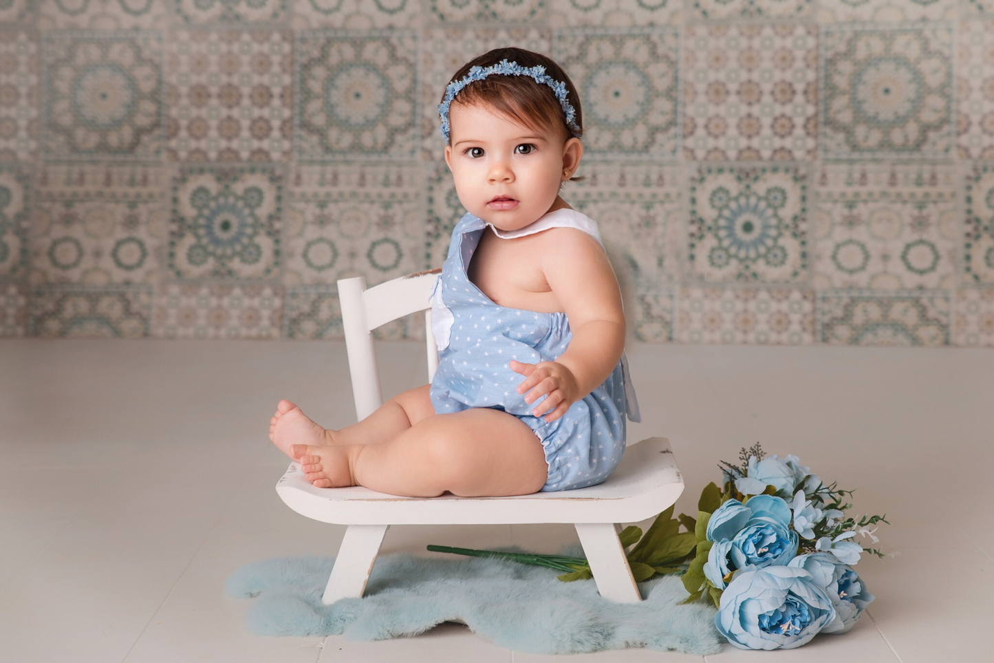 A curious baby sits comfortably on a white wide-base chair, dressed in a delicate blue polka-dotted dress, accessorized with a matching blue floral headband. The newborn photography prop is enhanced by a bouquet of silk blue flowers resting beside a soft, teal fur rug, all set against a backdrop of vintage-patterned tiles, creating a serene and charming scene.