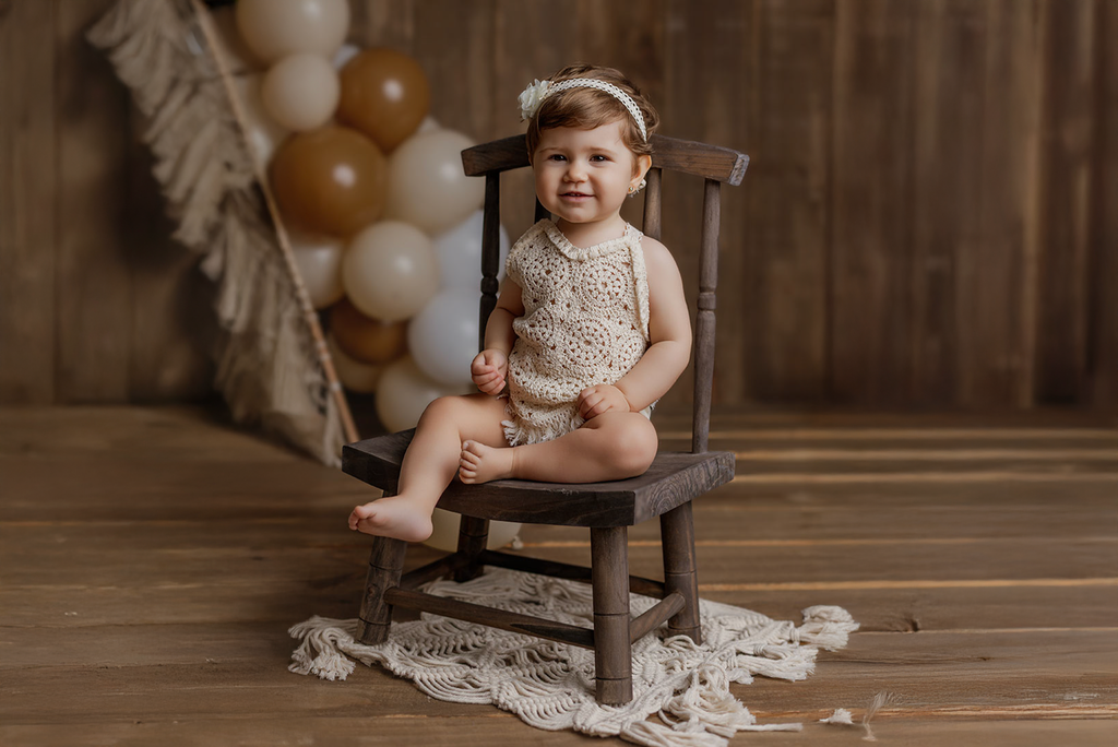 Baby sitting on a wooden chair with balloons in the background in baby photography setup