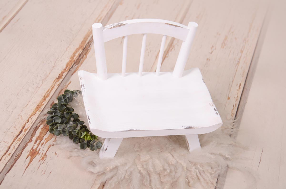 Wide base white newborn prop chair with distressing on a fur rug and greenery.