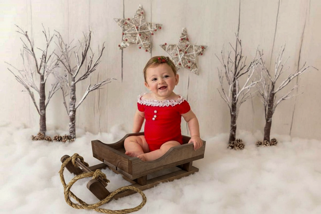 Smiling baby in red outfit sits on wooden sled newborn photography prop with snowy winter backdrop.
