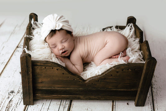 Newborn asleep in a rustic wooden bed, adorned with a white headband, highlighting the newborn photography prop.