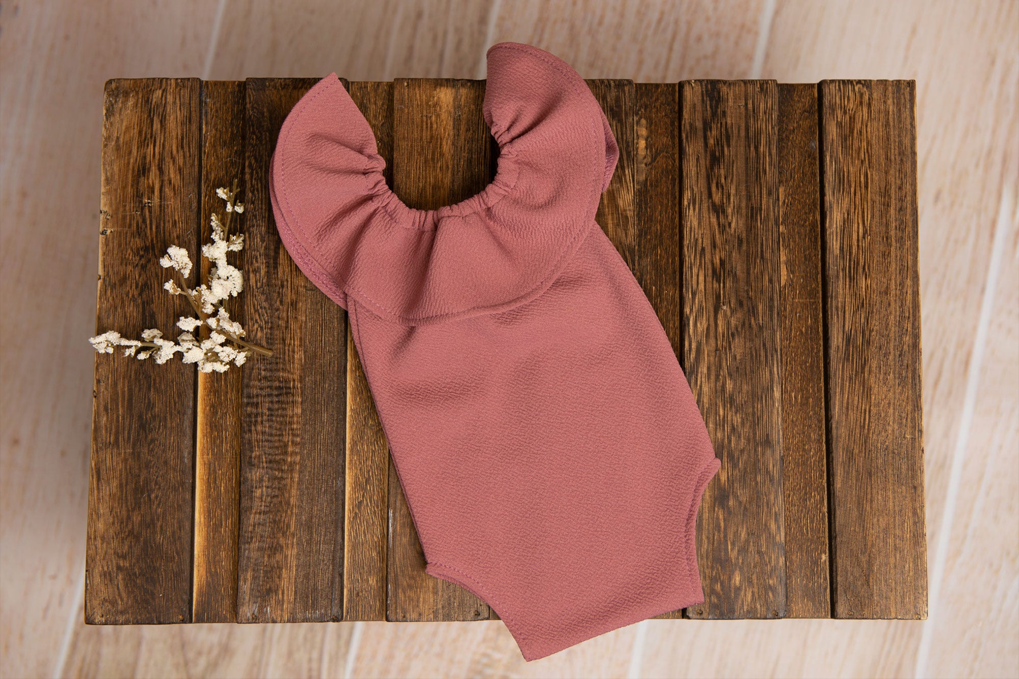 A mauve-colored ruffled sleeveless bodysuit for newborns laid on a wooden surface with a small bundle of flowers beside it.